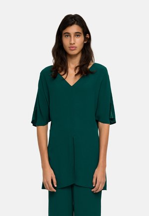 Młoda kobieta o ciemnych włosach, ubrana w luźną, ciemnozieloną tunikę z dekoltem w serek i rękawami do łokcia, stojąca na jednolitym tle.