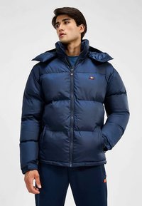 Marineblauwe donzen jas met een hoge kraag, ritssluiting en elastische manchetten. Heeft een logopatch op de borst en een gladde, glanzende textuur.