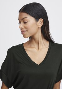 Mörkgrön V-ringad t-shirt med korta ärmar, tillverkad av ett slätt tyg. Har en avslappnad passform och lätt drapering runt axlarna.