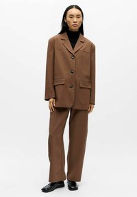 Bruine op maat gemaakte blazer met grote knopen, twee zakken aan de voorkant en revers met inkeping, gecombineerd met bijpassende wijde broekspijpen en zwarte schoenen.