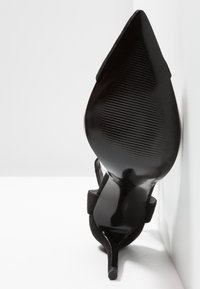 Chaussure à talon haut en daim noir avec bout pointu et talon fin, dotée d'une semelle texturée et d'une bride à la cheville, sur fond blanc.