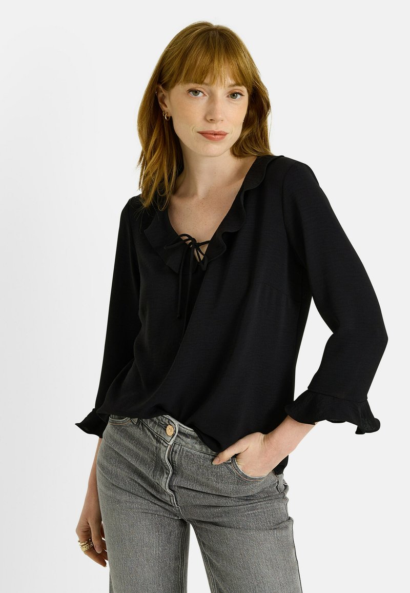 Blusa negra con volantes, escote en V y detallado con lazo, fabricada en un tejido ligero. Presenta mangas largas con puños rizados y un corte relajado.