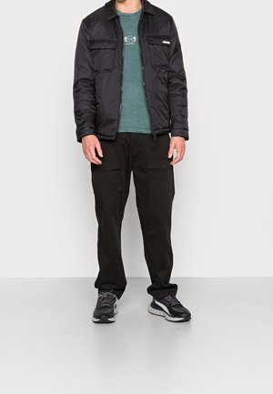 Man in een zwarte bomberjack over een groen graphic t-shirt, zwarte cargo-broeken en zwarte sneakers met witte zolen, staande op een grijze vloer.