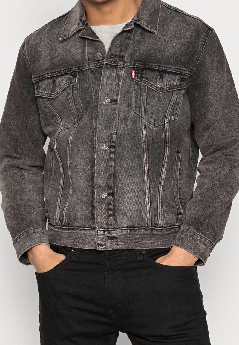 Homme portant une veste en jean noire délavée avec fermeture à boutons et poches poitrine, les mains dans les poches d'un pantalon noir.
