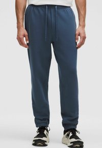 Man in marineblauwe joggers met elastische boorden en een trekkoord in de taille, gecombineerd met zwart-witte sneakers, staand tegen een effen achtergrond.