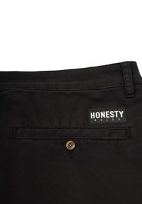 Schwarze Hosen aus strukturiertem Stoff, mit einer schmalen Gesäßtasche, einem braunen Knopf und einem Etikett mit der Aufschrift "EHRLICHKEIT REGIERT."