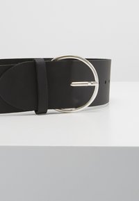 Ceinture en cuir noir avec une boucle circulaire en métal argenté, texture lisse, et trous ajustables pour personnaliser la taille.