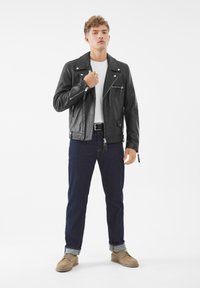 Junger Mann in einer schwarzen Lederjacke, weißem T-Shirt, dunkelblauen hochgekrempelten Jeans und beigen Schuhen, der vor einem weißen Hintergrund steht.