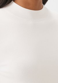Primer plano de una persona que lleva un suéter blanco de cuello cisne acanalado, mostrando la textura y el ajuste en la parte superior del pecho y el cuello.