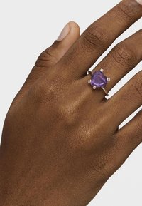 Anillo de gema en forma de corazón púrpura engastado en plata, con pequeños acentos. La banda es delgada con un acabado suave. Tono de piel visible.