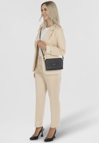 Schwarze Leder-Crossbody-Tasche mit strukturiertem Finish und einer rechteckigen Fronttasche, kombiniert mit einem beigen Anzug und hohen Schuhen.