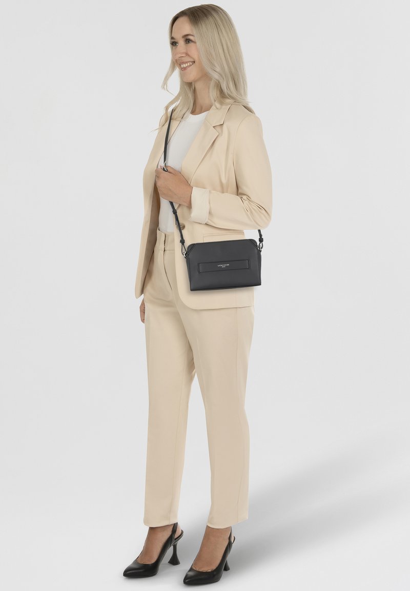 Schwarze Leder-Crossbody-Tasche mit strukturiertem Finish und einer rechteckigen Fronttasche, kombiniert mit einem beigen Anzug und hohen Schuhen.