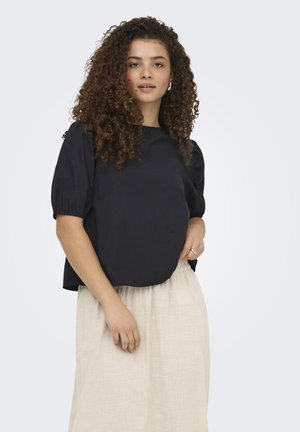 Junge Frau mit lockigem Haar, die eine schwarze Bluse mit kurzen Ärmeln und einen beigen Rock trägt, vor einem schlichten hellen Hintergrund stehend.