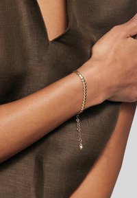 Złota bransoletka z sercowym wzorem i regulowanym łańcuchem, noszona na opalonej ręce. Tło to teksturowany, ciemnobrązowy materiał.