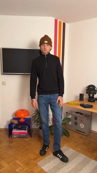 Svart zip-up hettegenser, blå jeans, svarte joggesko med hvite striper, og brun lue. Innendørs omgivelser med dekorative planter og møbler.