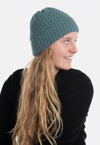 Gehaakte turquoise muts met een gestructureerde bovenkant en geribbelde rand, gemaakt van zacht materiaal. Draag met los, golvend blond haar en een zwarte top.