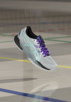 Hellgrauer Sportschuh mit mintfarbenen Streifen und lila Schnürsenkeln schwebt über einem Hallenboden mit farbigen Linien.