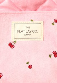 Tejido de forro polar rosa con patrones de cerezas bordados y una etiqueta que dice "THE FLAT LAY CO. LONDON." Textura suave, diseño acolchado.