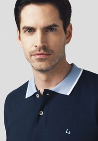 Granatowa koszulka polo z jasnoniebieskim kołnierzykiem i białymi wykończeniami, posiadająca dwa guziki oraz małe logo po lewej stronie. Gładka tekstura.