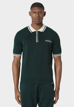 Mand iført en mørkegrøn poloshirt med korte ærmer, hvid krave og ærmestriber, stående mod en ensfarvet lys baggrund.