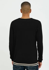 Maglietta nera a maniche lunghe con una texture a coste, caratterizzata da un collo rotondo e un orlo a strisce bianche in contrasto. La vista posteriore mostra maniche aderenti.