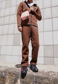 Person i brun träningsdräkt och svarta, röda och blå Nike sneakers, som håller en rosa Nike crossbody-väska, står på en betongplattform.