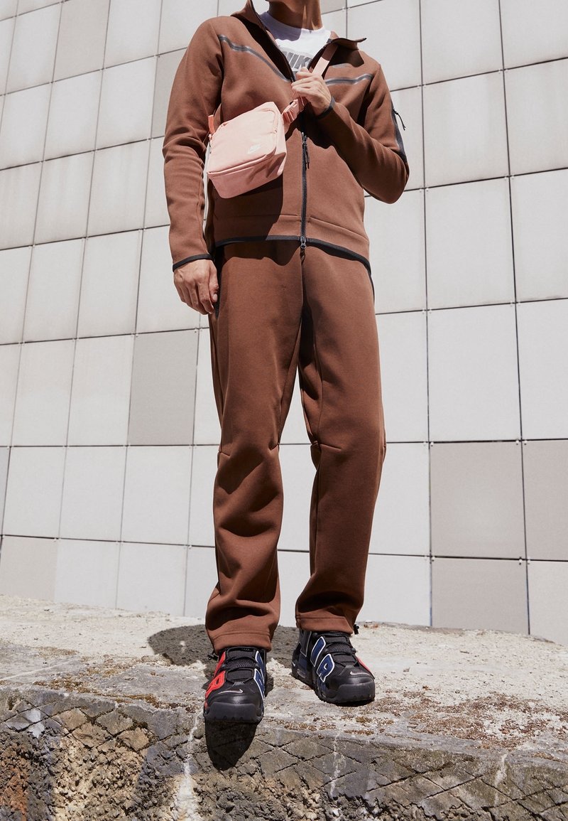 Person i brun träningsdräkt och svarta, röda och blå Nike sneakers, som håller en rosa Nike crossbody-väska, står på en betongplattform.