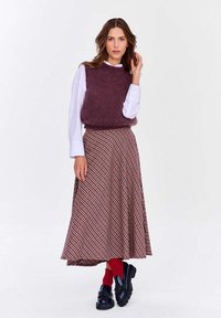 Pletená fialová bezrukávová sveter cez bielu košeľu, skombinovaná s voľnou červenou a čiernou kockovanou sukňou a čiernymi koženými topánkami.