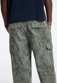 Cargo-Hosen im Tarnmuster, mit einer Mischung aus grünen und braunen Farbtönen, mehreren Taschen und einer entspannten Passform. Der Stoff wirkt langlebig.