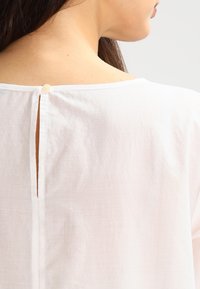 Vit linnetopp med sprund i ryggen och knappstängning, med en avslappnad passform och mjuk textur. Enkel, neutral design.