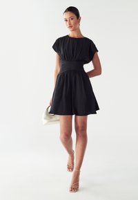 Woman in a short black dress with wide waistband, holding a white handbag, wearing nude strappy heels, standing against a plain light background.