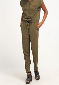 Une combinaison vert olive avec des manches courtes, un bouton à l'avant, une seule poche poitrine, une taille à cordon ajustable et des jambes fuselées. Portée avec des chaussures compensées noires.