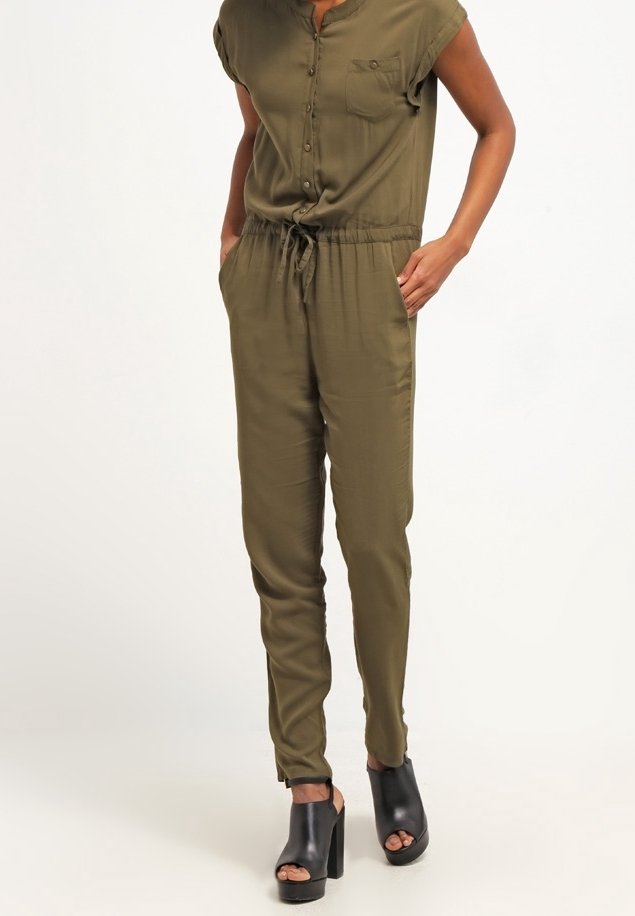 Une combinaison vert olive avec des manches courtes, un bouton à l'avant, une seule poche poitrine, une taille à cordon ajustable et des jambes fuselées. Portée avec des chaussures compensées noires.