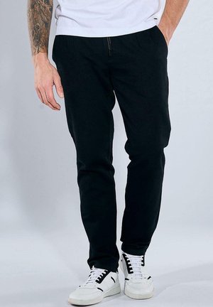 Black trousers with a smooth texture and tapered fit, featuring a zippered front pocket, paired with white sneakers with black accents.