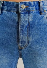 Blaue Jeans aus Denim mit einer ausgeblichenen Waschung, ausgestattet mit einem Knopfverschluss, Kupfernähten und Gürtelschlaufen. Glatte Textur mit subtiler Schattierung.