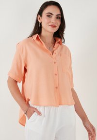Short-sleeve, coral button-up shirt with a collar, single chest pocket, relaxed fit, and side slits. Made from lightweight fabric.