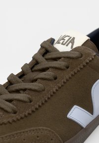 Baskets en cuir suédé marron avec des lacets ton sur ton, un logo en caoutchouc blanc contrastant et des détails cousus. Présente une surface texturée et une forme arrondie.