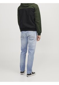 Giacca con cappuccio con pannelli verdi e neri, orlo elasticizzato; jeans di denim azzurro con dettagli di cuciture sottili; scarpe da ginnastica nere con suole bianche.