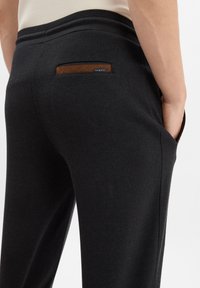 Black trousers made from woven fabric, featuring a rear pocket with a brown leather accent and a small brand tag. Elastic waistband design.