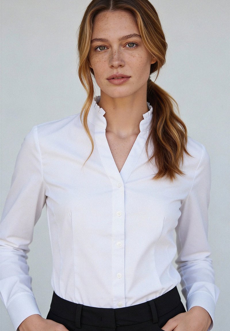 Jeune femme avec de légères taches de rousseur et de longs cheveux ondulés attachés, portant une chemise blanche à boutons avec un col à volants, sur un fond uni.