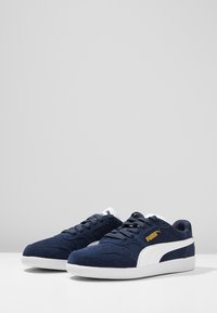 Marineblaue Wildleder-Sneaker mit weißer Gummisohle, ausgestattet mit einem weißen Seitenstreifen und goldenem Logo. Schnürdesign mit gepolstertem Kragen für zusätzlichen Komfort.