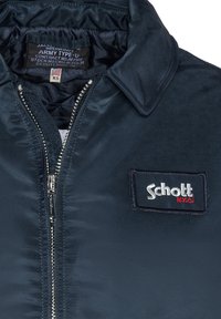 Námořnicky modrá bomber bunda se lesklým saténovým povrchem. Obsahuje kovový zip, klopový límec a nášivku s označením "Schott N.Y.C.".
