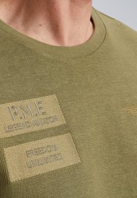 Maslinasto zelena pletena majica s okruglim izrezom, sa dva vezena zakrpa: "P.M.E. LEGEND AVIATOR" i "FREEDOM UNLIMITED."