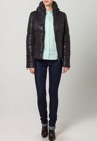 Giacca a vento nera con finitura lucida, chiusura con zip e design trapuntato. Indossata sopra una camicia verde chiaro e jeans skinny scuri.