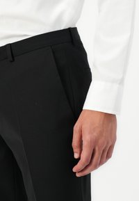 Pantalon noir ajusté en tissu lisse, avec des plis avant, une taille cintrée et des poches latérales. Main visible près de la taille.