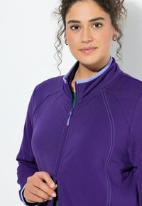 Fioletowa kurtka na zamek błyskawiczny z kontrastującym jasnoniebieskim kołnierzem. Charakteryzuje się teksturowaną tkaniną, eleganckim designem oraz widocznymi detalami szwów.