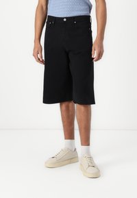 Black cotton bermuda shorts with a loose fit, five pockets, and silver hardware. Paired with light sneakers and white socks.
