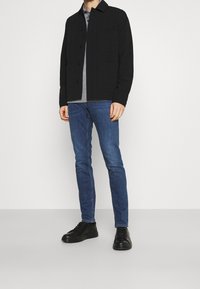 Černá bundička na zapínání s klasickým límcem, pod ní šedé tričko, modré džíny s úzkým střihem a černé tenisky. Hladká textura, neformální design.