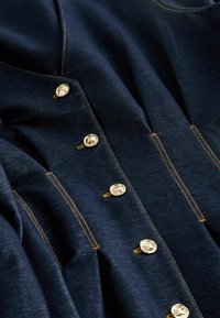 Tmavomodrá rifľová bunda so zlatými kovovými gombíkmi, dvojitými vreckami a kontrastným žltým prešívaním. Mäkká textúra so štruktúrovaným dizajnom.