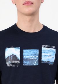 Nærbilde av en person som har på seg en marineblå skjorte med tre hav-tema grafiske firkanter og tekst inkludert "TOM TAILOR."